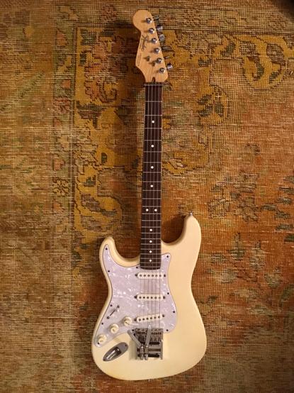 White Stratocaster electric guitar. Bone white body, white pickguard. Dark rosewood fretboard. Lying on a yellow and orange overdyed Persian rug.