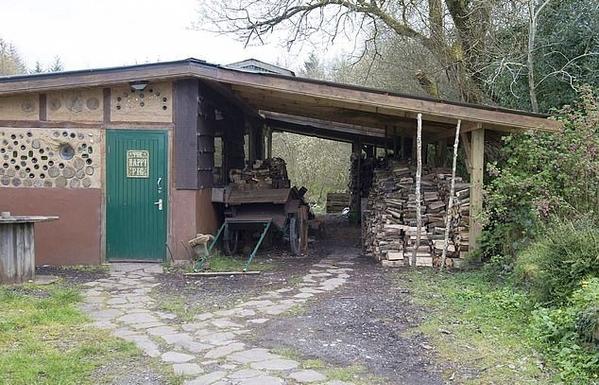 A photograph of the outside of The Happy Pig, Co. Galway, Ireland.

A path leads to the door of the wood/cob structure, featuring door sign "The Happy Pig", and outside a cart parked up, large woodpile and rural wooded backdrop.