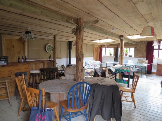 Inside the main space of the Happy Pig.  The bar, a wood burning stove, chairs, tables , sofas, table football and table tennis visible inside the wood/cob structure.
