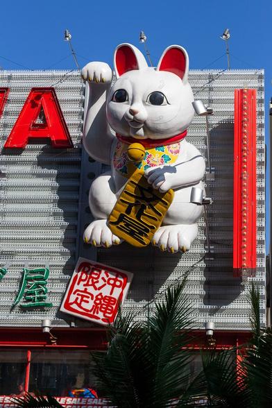 A huge Maneki-neko on the side of a building. Maneki-neko is the Japanese beckoning or lucky cat symbol; a white cat standing on its back legs with one paw raised, waving. This one is several metres high and on the flat surface of the upper part of a commercial property on a high street. Its ears are red, it is wearing a red collar with yellow circular bell, and what looks like a floral, patterned scarf. In its non-raised paw it is holding a yellow sheet with fan folds on it and Japanese writing in black. The building side almost looks corrugated and has some Japanese characters below the cat as well as the letter A, large and in red, to the left. Below the corrugated section of the building are some windows at street level, in shade from everything above, and there is a fern-like tree. It's a bright, sunny day with clear blue sky behind the cat's ears that extend above the building's roof.