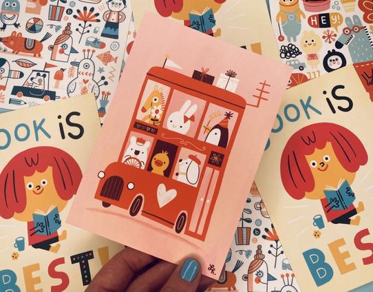 Linzie hand holding a postcard featuring a red bus full of cute characters on a pink background.