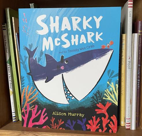 Picture of picture book on bookshelf the cover has a smiley shark with a crab on her nose. The title of the book is Sharky McShark and the Teensy Wee Crab!