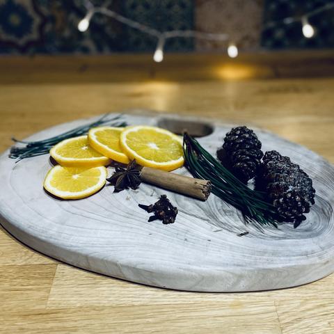 A wooden board on a kitchen counter with pine needles and cones, sliced orange, star anise, a cinnamon stick and cloves arranged on it. Some fairy lights hang in front of the tiles in the background.