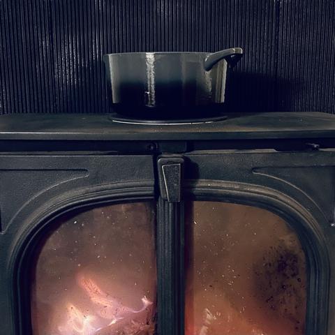 A grey pot sits on top of a lit wood burner; the sooty windows show the orange glow of the fire within