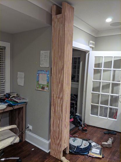 a messy room with a half-finished wooden tower in the middle of it. The tower is plywood, and the tower is roughly 9 inches on each side. There's a hole on the bottom and the very top for cats to go in and out. The tower just about touches the ceiling (8ft tall).