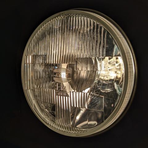 Koito round headlamp illuminated at night on low beam setting. Image exposure and contrast yielded a black background with just the headlamp illuminated.

The headlamp has vertical patterns on the top third and bottom third of the disk. The middle third has lens patterns to properly shape the light emitted.

The center of the lens has the stylized Koito logo and H4 below the logo.