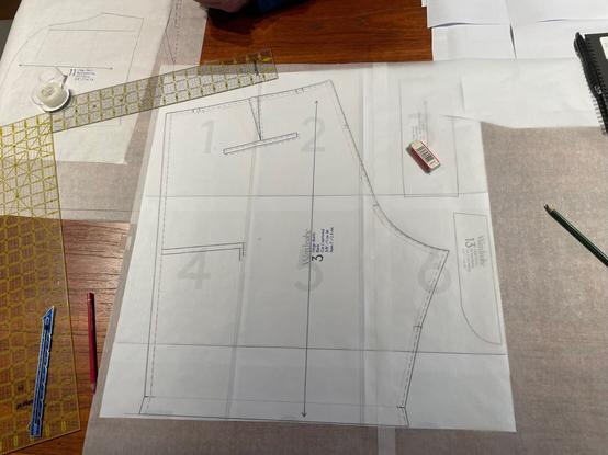 a reddish-brown table seen from above.  the surface is cluttered with pattern drafting implements.  a flat pattern piece for the rear of a pair of shorts lies in the middle of the photo, overlaid with tracing paper.  to the right of the pattern piece, a plastic eraser and a green colored pencil are visible.  to the left, two quilting rulers, a roll of cellphane tape, a sewing gauge, and a red colored pencil is seen.  other pattern pieces and tracing paper are scattered in the upper corners of photo.