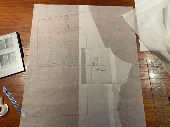 the surface of a reddish wood table, with a back shorts pattern piece laid out in the center.  the curve of the crotch points to the right of the photo.  a section of the pattern along the crotch line has been cut out and shifted 1" to the right.  a quilting ruler and pieces of paper are seen in along the right edge of the photo.  along the left of the photo, a tablet, a sewing gauge, and a roll of cellophane tape can be seen.