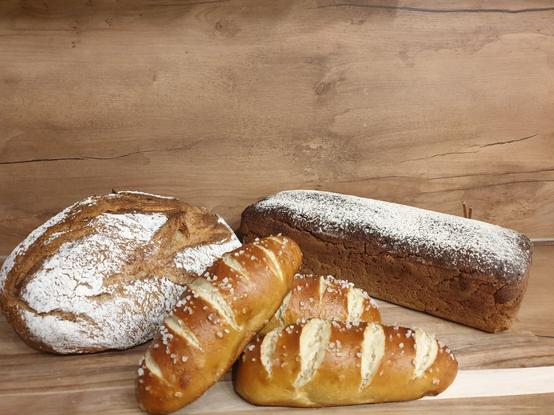 Im Vordergrund liegen 3 Dinkellaugenstangen. Schräg rechts dahinter ein Roggenschrotbrot mit Dinkel in Kastenform gebacken. Hinten links ein frei geschoben gebackenes Dinkelsauerteigbrot aus Ruchmehl