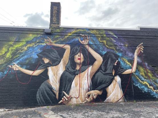 A large mural on a brick building of three women as The Fates, holding a long red string in their hands.