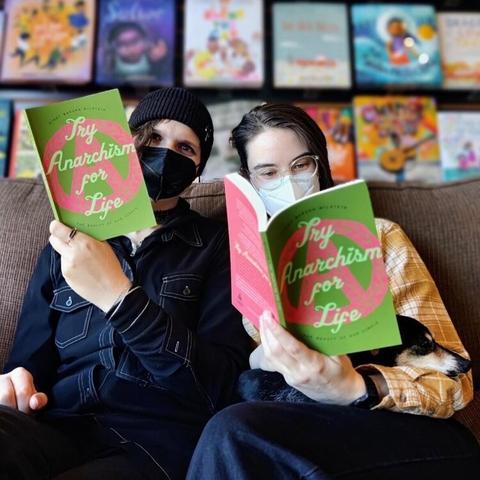 Two people and a small dog sitting on a couch reading my latest book, with the cover visible, with its two tones of green and big pink circle A