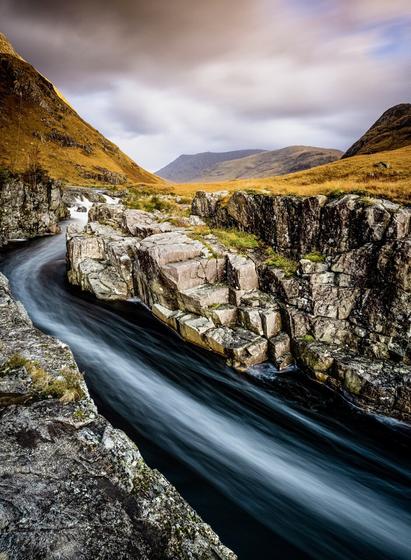 A cold day in autumn as the sun casts an orange glow on the landscape of Glen Etive. Tall mountains surround the scene with blurred clouds covering the sky. A gorge in slick black rock contain a fast flowing river after heavy rainfall. Bubble formed by a waterfall further upstream float by. As this is a long exposure image, the bubbles form long white streaks in the darkness of the river creating a sense of speed.