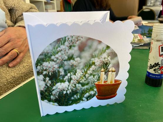 White greetings card with magazine images of white Heather flowers and candles on the front, standing on a green table with an older white lady’s hand seen behind
