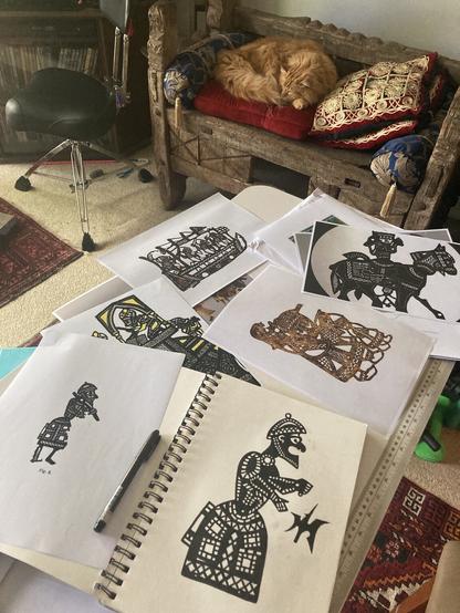 A collection of Egyptian shadow puppets are printed and scattered on a table, one is a drawing in a sketchbook. In the background a ginger cat sleeps comfortably.