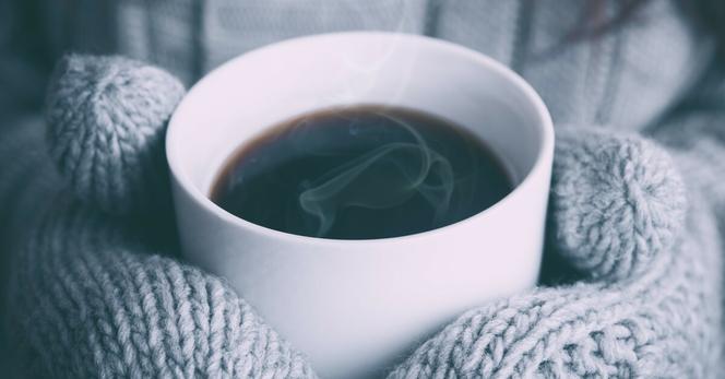 Hands covered in light grey, knit mittens clutch a steaming mug of coffee. Image by Alex Padurariu on Unsplash