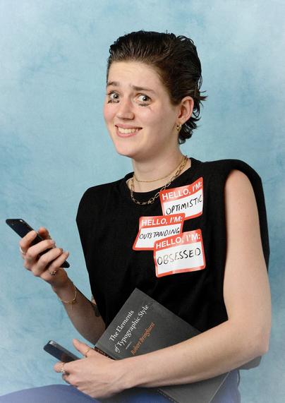 Photo of a young person holding two phones, a copy of “The Elements of Typographic Style” book, and wearing 3 generic name tags saying "Hello I'm optimistic”, "Hello I'm outstanding” and "Hello I'm obsessed”.