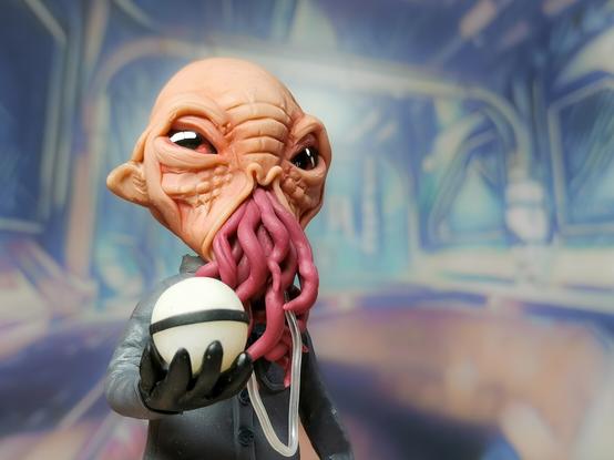 Small handmade clay figure of an alien species called an Ood, from TV's Doctor Who. It has big eyes, pointy ears, bald head and long worm like things where it's mouth should be. It wears a grey suit and holds a ball in it's hand which it uses to communicate.