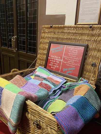 An open wicker hamper filled with brightly coloured knitted blankets. A sign on the inside of the hamper lid reads ‘Welcome to St Anne’s. Feel free to borrow a blanket to help you stay cosy while you’re here. God loves you!’