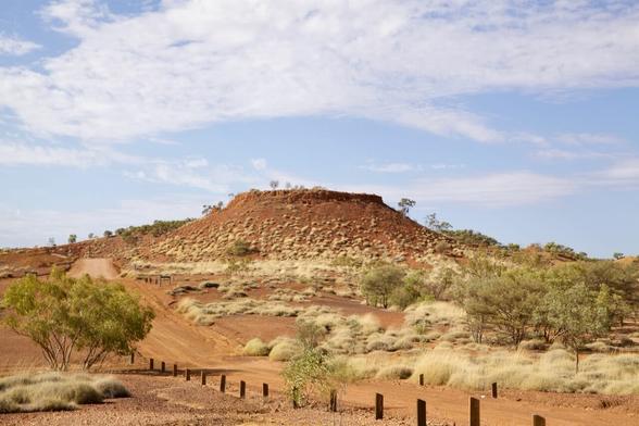 A large hill with a flat top is the centre of the photo. A dirt road runs past it into the distance. There are clumps of spinifex grass dotted on the sloped sides of the hill and the plain leading to it
