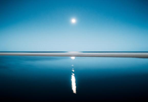 Blue sky with the moon centered,  below it a band of ocean with the moonlight shining in the centre, below that a sandbar and below that the lagoon with the moon's reflection stretching down the middle