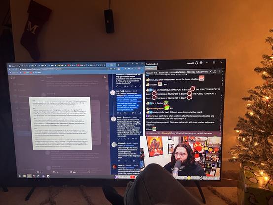 Flat screen TV with Twitch stream on, solid background, stocking with a letter M, and Christmas tree to the right side of the photo.