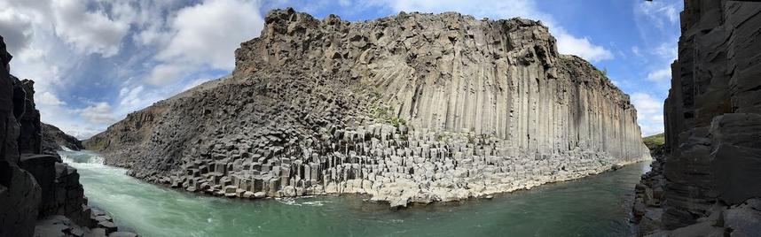 A panoramic photo of Studlagil canyon in Iceland, with a partially cloudy sky, grey basalt columns, and light green water.  A waterfall can be seen on the left side in the distance.