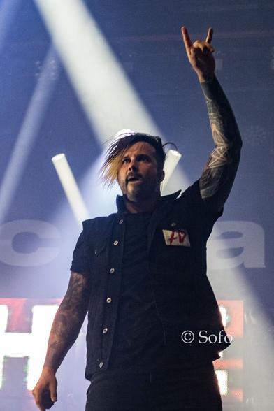 The photo shows Electric Callboy's shouter Kevin Ratajczak at a concert. One of his hands is lifted to show a metal fork (the hand gesture used by metalheads). His hair is mostly dark brown, but the tops of the longer strands of his bangs are dyed blonde.
