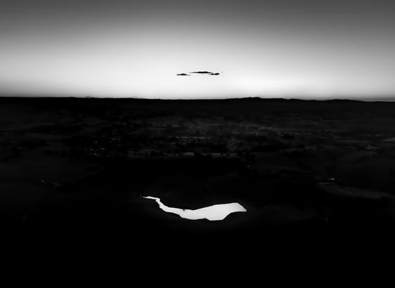 Graphic monochrome image: white sky with lone dark cloud. Black ground with a single white small lake mirroring the shadow of the cloud
