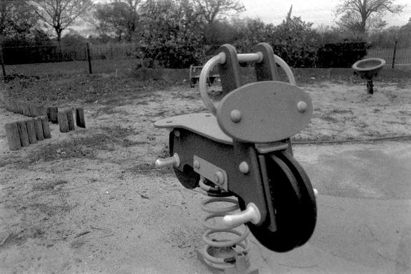 A black and white film photograph of a piece of playground equipment for younger children in the form of a wooden motorbike shaped seat on a large metal spring.