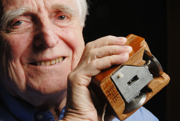 A photo of an aged Doug Englebart, smiling and holding the prototype of the first computer mouse.