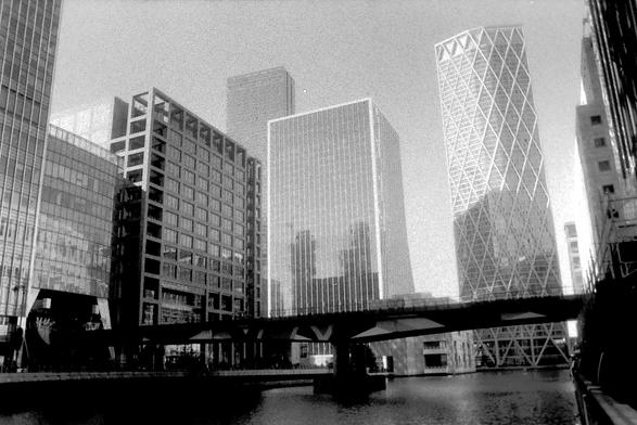 A black and white film photograph towering skyscrapers surrounding a body of water at Canary Wharf, London.