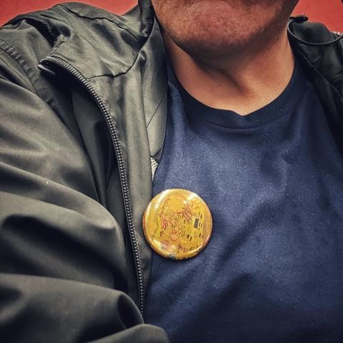 Acercamiento al torso de un hombre vestido de chamarra y camiseta. Sobre ésta está fijado un botón metálico decorado.