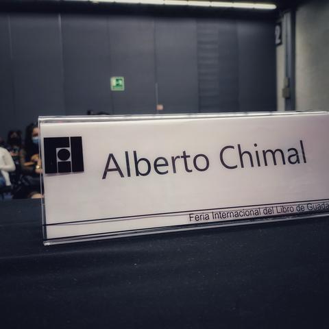 Sobre una mesa en un auditorio, un personalizador de plástico en el que se puede ver el logotipo de la FIL Guadalajara y el nombre de Alberto Chimal.