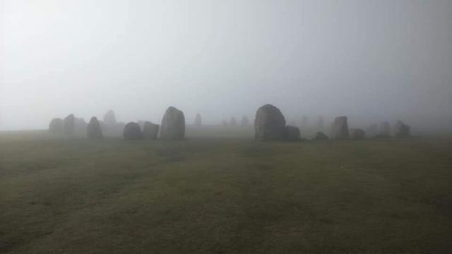 A foggy scene. The stone circle is viewed from the side with two larger, entrance,  stones and a gap in between prominent. There are about 30 stones that make up the circle.