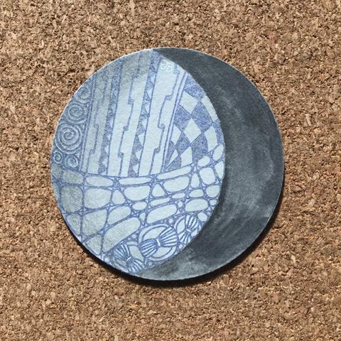 A grey Zendala Zentangle tile that looks like a waning moon with a slice coloured in solid black. The other remaining area is filled with various patterns drawn in a blue silver metallic pen and coloured over with graphite so that is is shiny and reflective