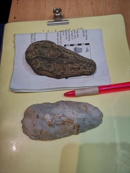 Two neolithic tools on a yellow card. Top One is made of Andesite from the Cheviot Hills, the other is flint and found on the Isle of Wight. A red pen is in between them.