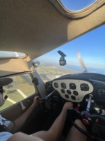 A pilot at the controls of a small aeroplane with green farmland and blue ocean in the distance.