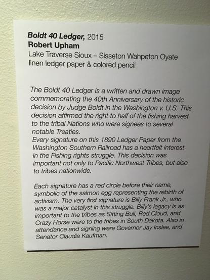 Sign that reads: 
Boldt 40 Ledge, 2015
Robert Upham
Lake Traverse Sioux -- Sisseton Wahpeton Oyate 
linen ledger paper & colored pencil

The Boldt 40 Ledger is a written and drawn image commemorating the 40th Anniversary of the historic decision by Judge Boldt in the Washington v US. This decision affirmed the right to half of the fishing harvest to the tribal Nations who were signees to several notable Treaties.
Every signature on this 1890 Ledger Paper from the Washington Southern Railroad has a heartfelt interest in the Fishing Rights Struggle. This decision was important not only to the Pacific NW Tribes, but also nationwide.
Each signature has a red circle before their name, symbolic of the salmon egg representing the rebirth of activism. The very first signature is Billy Frank Jr, who was a major catalyst in this struggle. Billy's legacy is as important to the tribes as Sitting Bull, Red Cloud, and Crazy Horse were to the tribes in S Dakota. Also in attendance were Gov Jay Inslee and Senator Claudia Kaufman.