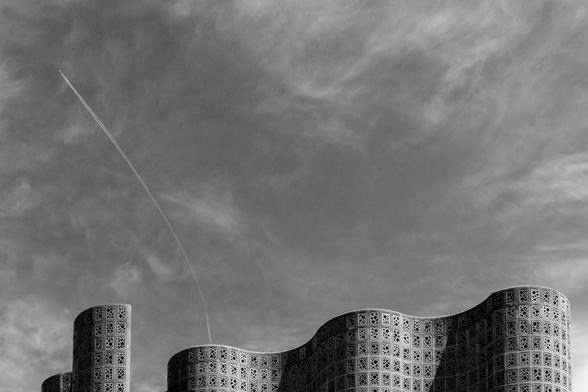 Black and white photograph of the curved, undulating top of the 1964 New York World Fair science pavilion, now the New York Hall of Science museum.  There are oddly shaped dark glass “windows” in the structure as it rises up, and the shape of the structure causes alternating light and shadowed areas. Some clouds are visible, and an aircraft is in the top left, leaving a curved contrail that leads back to the top of the building.