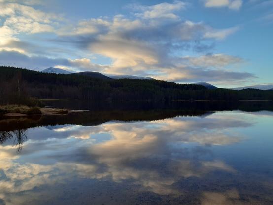 Row of forested hills in front of a distant set of snow-capped mountains. In front of the hills is a still loch reflecting a blue sky with dotted fluffy clouds (also visible in the sky above)