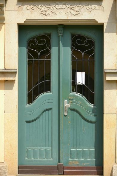 Photo of a green double doors with windows. There is an ornamental grille on each window. The right side door has a white notice stuck to the window from the inside. There is a decorative stone carving above the doors.