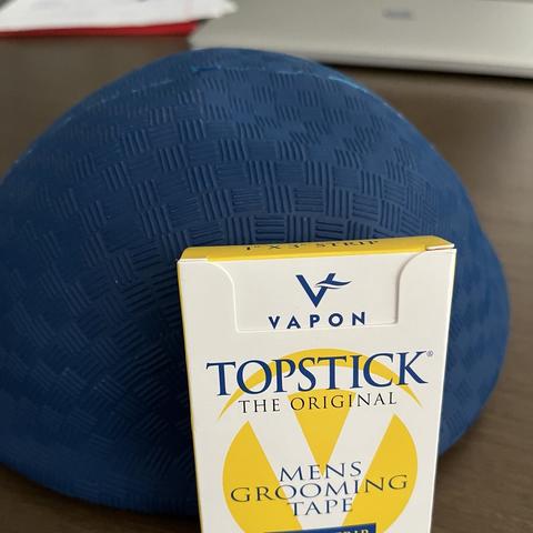 A blue uninflected dodgeball and a package of TOPSTICK brand toupee adhesive on a table in the foreground; laptop and notebook in the background.