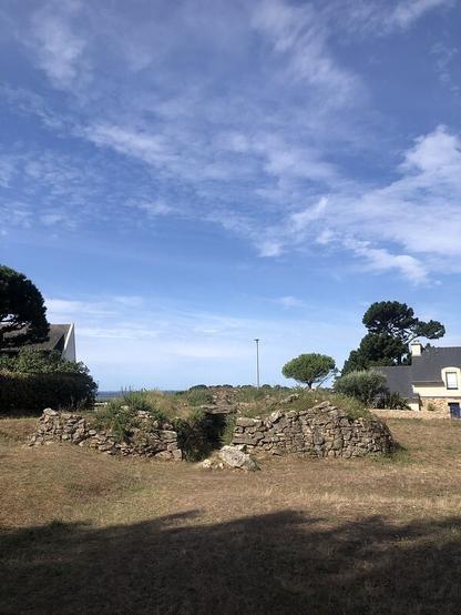 A beautiful sunny day. The sky is blue with wispy clouds, the sun is shining. A burial chamber with drystone walling and quite a lot of gorse growing on it. It looks kind of slumped - the mound has been removed. Youc an see one of the capstones within the passage. Behind it there are some good pine trees, Scots pine I think (or French equivalent) and on either side of the picture, the roofs of houses. The house on the right also has windows visible. You can just see the sea, too, on the horizon.