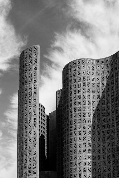 Black and white photo of the tall, undulating walls of the New York Hall of Science museum, the former 1964 Worlds Fair science pavilion. The space between the outer wall and the epically of the main hall form a valley, partially in shadow. There are clouds visible, and the expanse of wall are peppered with the irregular shapes of the dark window clusters embedded in tiles across all surfaces.