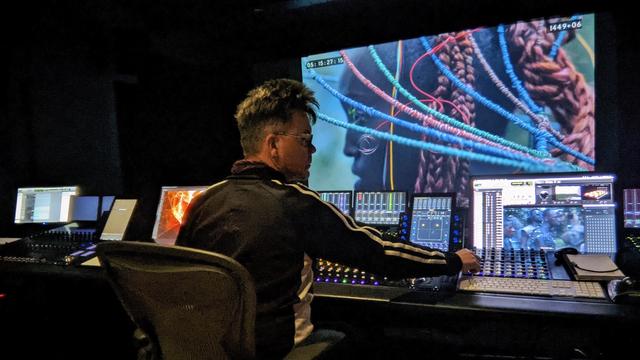 A white guy with glasses twiddles a knob on an extremely high-tech console with many data panels and LEDs. On a big movie screen in the front of the room is a striking image of a beautiful black woman with ornate red braids and swooping complex head gear