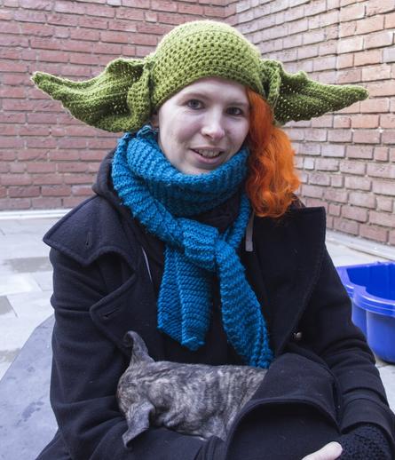 Ein gestromter Sloughi Welpe schläft auf dem Schoß von Lara, die einen Mantel trägt und eine grüne Mütze mit Yoda Ohren auf dem Kopf trägt. Die Ohren des Welpens ähneln optisch den Ohren der Mütze.
