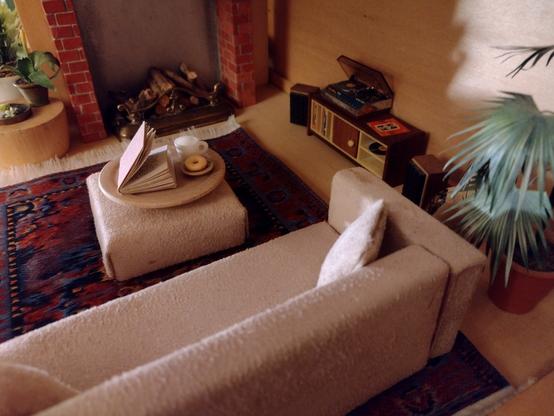 A photograph of a dolls house livingroom where the scene is set for sitting comfortably in front of an open fire. The fire surround is natural brick. Sunlight strikes the back wall, which is bare wood. The only floor covering is a large oriental rug in dark burgundy. A large houseplant stands in the corner. The sofa is modern and cream coloured with square lines. Along the wall is a nineteen sixties style music cabinet and a record player with the lid open. On a large square footstool is a tray with a book, a cup of tea and a doughnut.