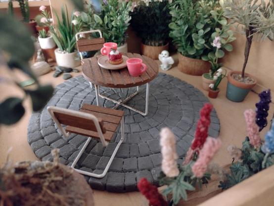 A photograph of a dolls house roof terrace. On a circular paved patio sit a garden table and two chairs. They are modern, slatted wood with white metal legs. The table is set for tea and cake for two. Surrounding the patio are a variety of different flowering plants and shrubs in pots.