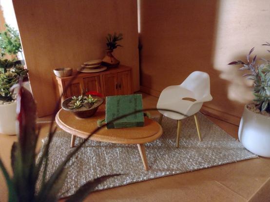 Photograph of a dolls house study. The only floor covering is a jute rug. A white mid century modern plastic chair sits in front of a kidney shaped wooden table. On the table are a book and a large bowl planted with succulents. The book is The Young King by Oscar Wilde. There is a modern oak sideboard along one wall. Sunlight strikes the back wall, also illuminating the leaves of a tall houseplant in a large white ceramic planter. All is peace.