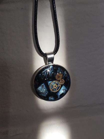 Steampunk-themed pendant A. It has a round 24mm silver bezel pendant  inset with polymer clay background. Two vintage watch mini cogs float on top of the background, and the pendant is set under resin. The background is a light iridescent metallic blue, with a partial print of a dark copper cog. The pendant hangs from a waxed cotton cord in a shaft of late afternoon sunlight against an off white brick wall.